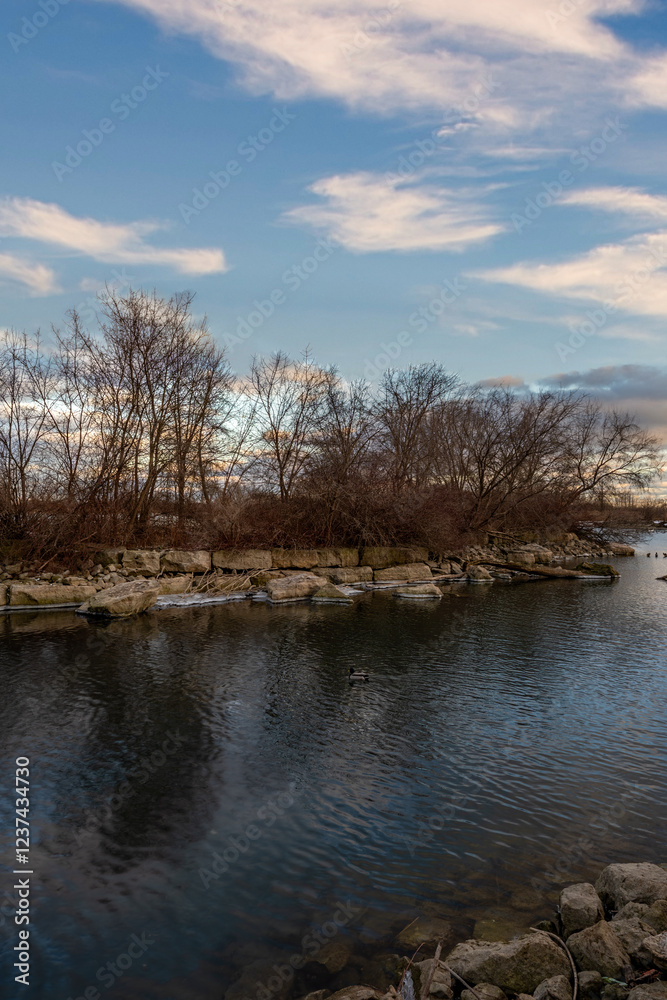 Winter scene: calm water reflects a sky, barren trees line a rocky shore. Winter scene: calm water reflects a sky, barren trees line a rocky shore.