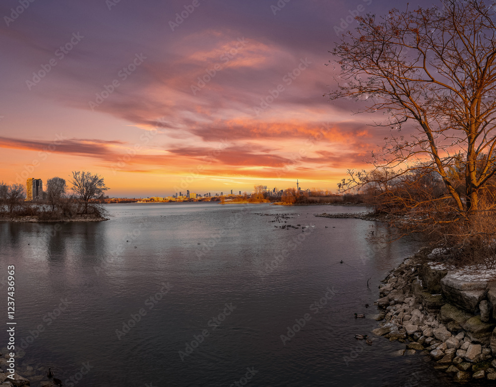 Sunset cityscape reflected on tranquil waters, framed by winter trees. Sunset cityscape reflected on tranquil waters, framed by winter trees.