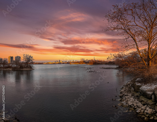 Sunset cityscape reflected on tranquil waters, framed by winter trees.