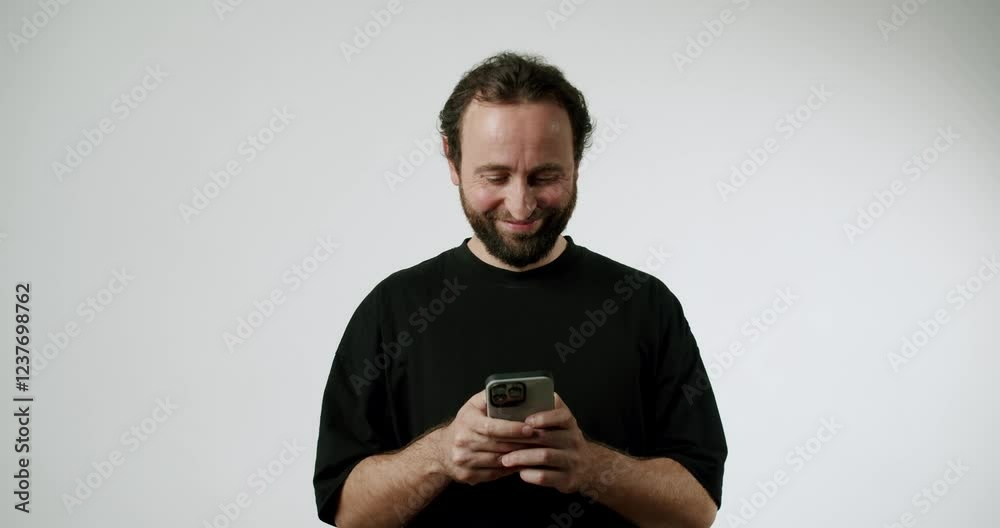 A man with a beard, wearing a black t-shirt, stands against a white background looking at his phone. He starts with a serious look, but then breaks into a smile as if reading a message. A man with a beard, wearing a black t-shirt, stands against a white background looking at his phone. He starts with a serious look, but then breaks into a smile as if reading a message.
