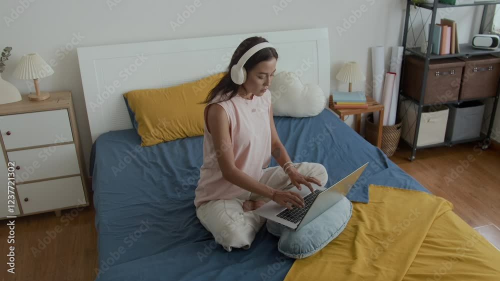 View from above of young Caucasian female freelancer wearing wireless headphones typing on laptop in cozy bedroom View from above of young Caucasian female freelancer wearing wireless headphones typing on laptop in cozy bedroom