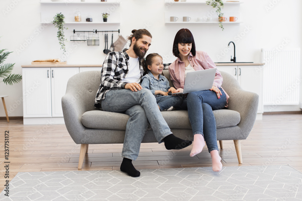 © sofiko14 - White family with mother, father, and young girl sitting on sofa watching laptop. Casual, joyful setting in modern living room. Happy, relaxed atmosphere with technology and bonding.