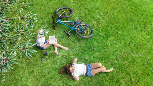 Young girls with bicycle in park, listening music with headphones and reading book, two student girls study and relax outdoors sitting on grass near bike, aerial drone view from above