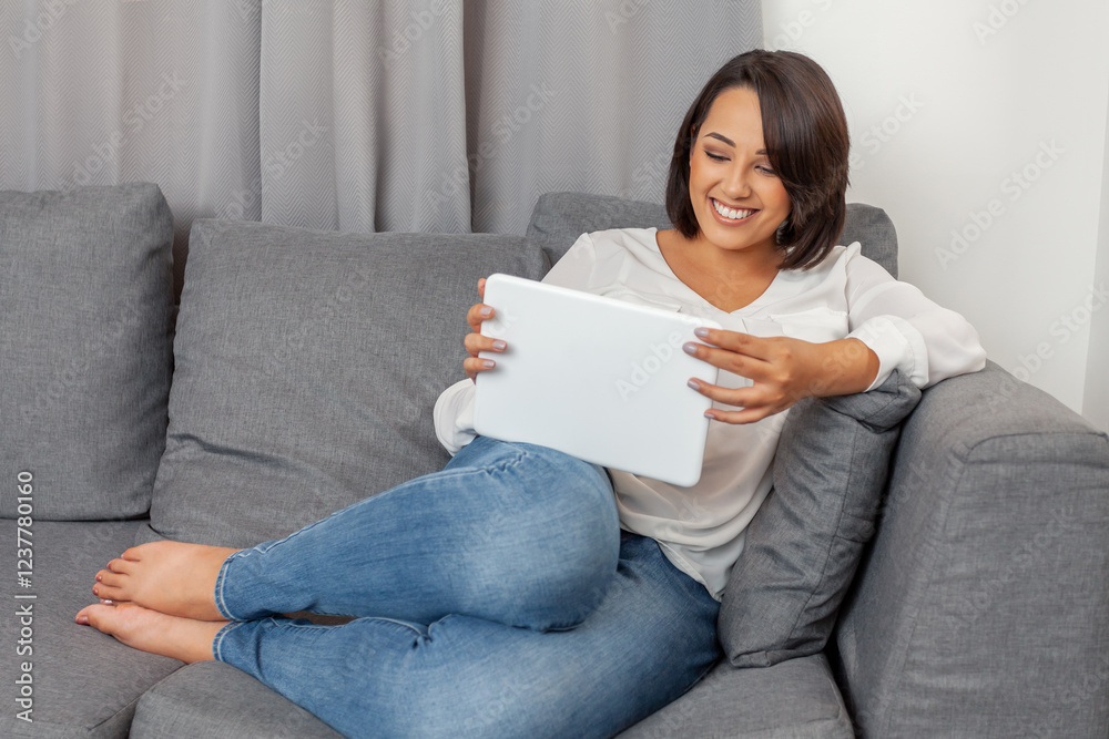 Young Asian Woman Relaxing on Grey Couch Using Digital Tablet, Wearing Casual Clothes and Enjoying Entertainment at Home Young Asian Woman Relaxing on Grey Couch Using Digital Tablet, Wearing Casual Clothes and Enjoying Entertainment at Home