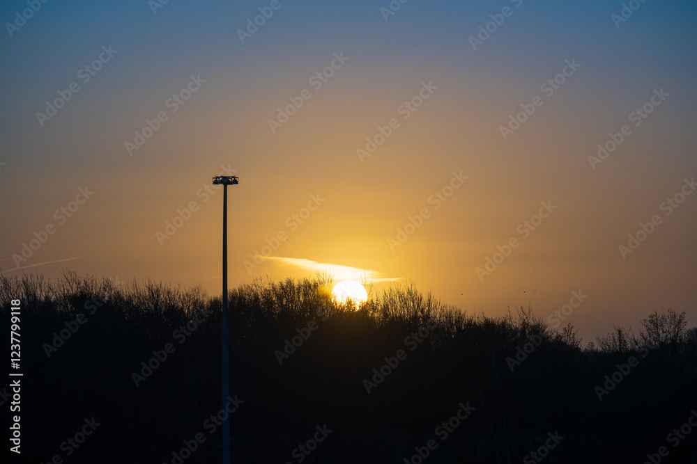 Fototapeta premium Dramatic winter sunrise behind silhouetted treeline with tall floodlight pole in the foreground. Golden morning sun breaking through the dark sky creates an atmospheric British landscape