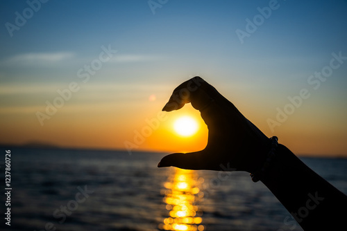 silhouetted hand forming a half-heart gesture in front of a glowing sunset over the ocean photo