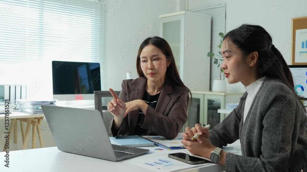 Two young businesswomen are actively engaging in a discussion, utilizing a laptop and analyzing documents, within a contemporary office environment Two young businesswomen are actively engaging in a discussion, utilizing a laptop and analyzing documents, within a contemporary office environment