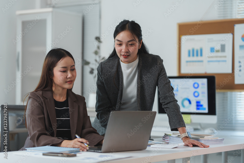 Two asian businesswomen are collaborating on a project, using a laptop in their modern office, surrounded by paperwork and charts displayed on a nearby corkboard and computer monitor Two asian businesswomen are collaborating on a project, using a laptop in their modern office, surrounded by paperwork and charts displayed on a nearby corkboard and computer monitor