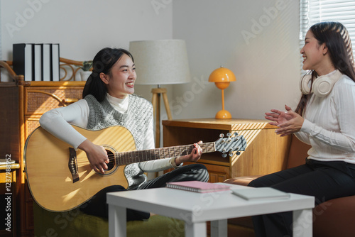 Two young asian women are enjoying their free time, playing music together in their living room, with one playing an acoustic guitar and the other singing along photo