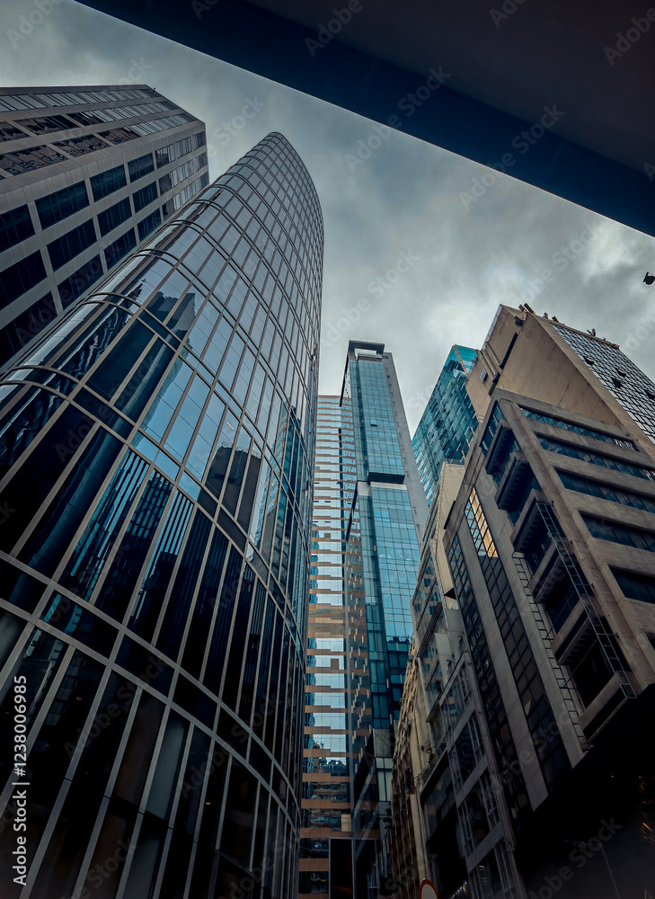 A low-angle, close-up view of skyscrapers in Hong Kong, their glass facades reflecting a cloudy sky. The buildings rise dramatically, creating a sense of towering urban density. A low-angle, close-up view of skyscrapers in Hong Kong, their glass facades reflecting a cloudy sky. The buildings rise dramatically, creating a sense of towering urban density.