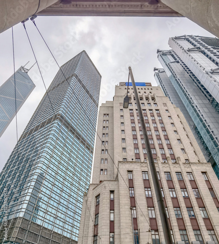 Low-angle close-up view of skyscrapers in Hong Kong, showcasing a mix of modern glass towers and older colonial-style architecture under an overcast sky.  The image captures the city's architectural c photo