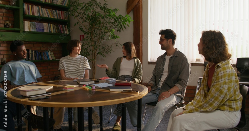 Obraz premium A group of young people are smiling and talking while sitting around a round table indoors. Notebooks and books on the table. Daylight