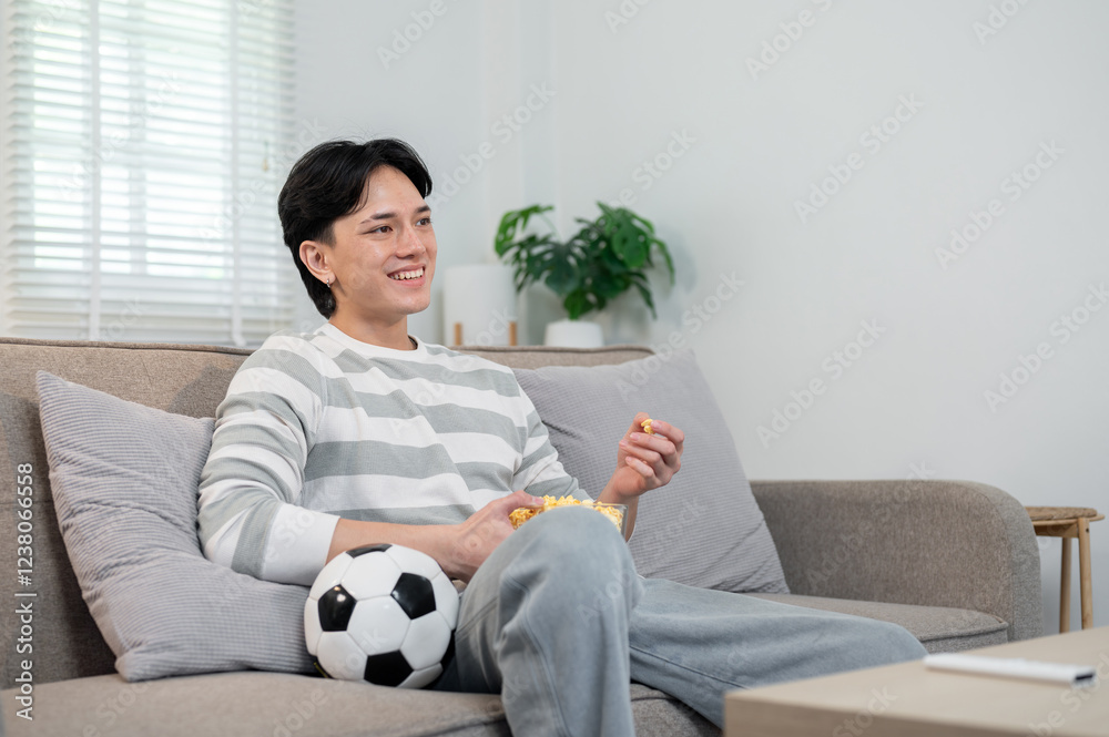 A relaxed Asian man enjoys eating popcorn while watching his favorite football team's match on TV. A relaxed Asian man enjoys eating popcorn while watching his favorite football team's match on TV.