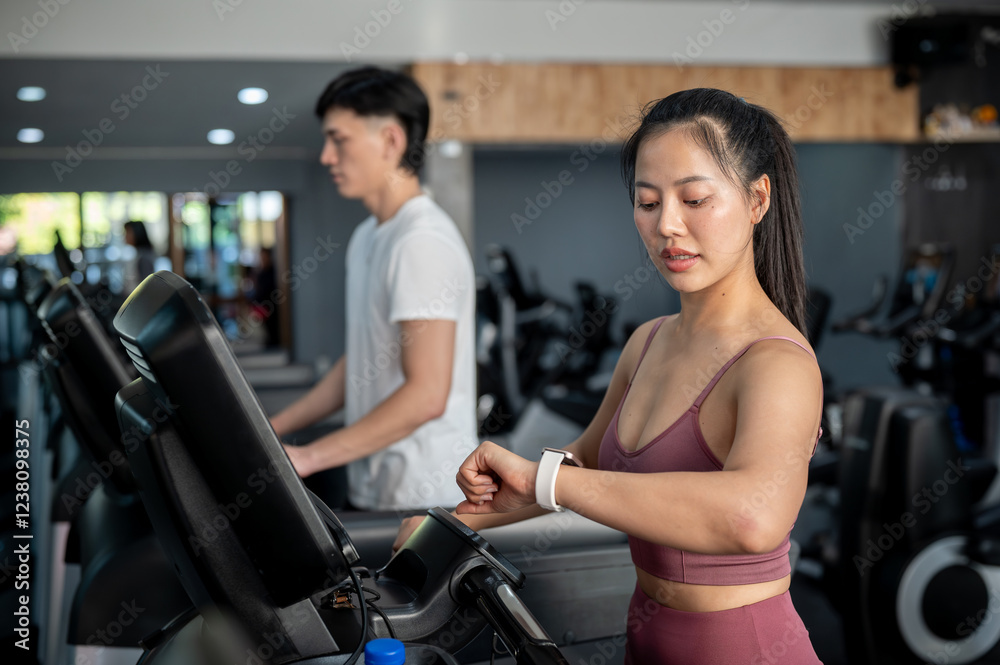 A sporty Asian woman is checking her calorie count on her smartwatch while walking on a treadmill. A sporty Asian woman is checking her calorie count on her smartwatch while walking on a treadmill.