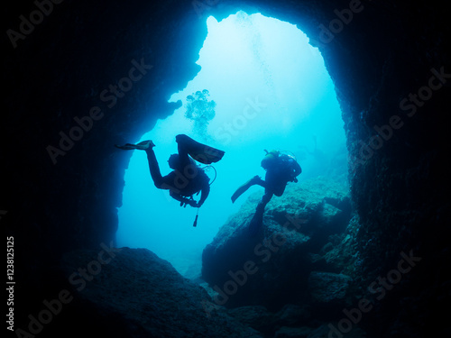 silhouette of two unrecognizable scuba divers diving in dark cave with stony formations undersea in deep clear water, concept of active lifestyle at nature and underwater adventure, copyspace for text