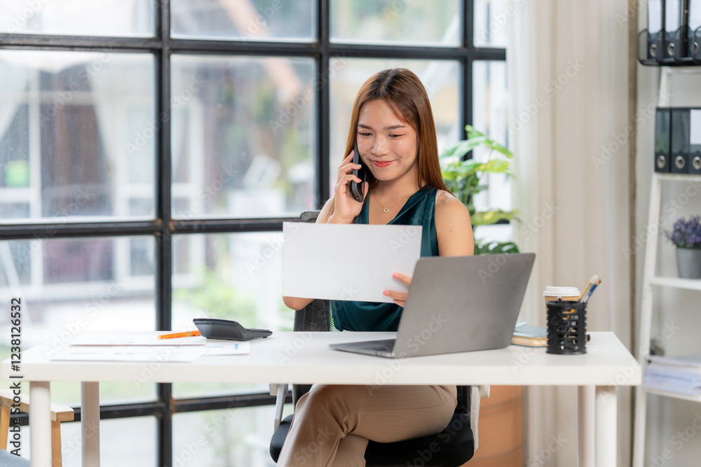 Focused Businesswoman: A smiling young businesswoman sits at her modern desk, efficiently managing her workload. She's on a phone call, reviewing documents, and using her laptop. Focused Businesswoman: A smiling young businesswoman sits at her modern desk, efficiently managing her workload. She's on a phone call, reviewing documents, and using her laptop.
