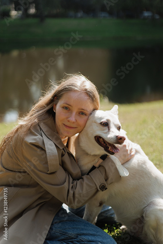 young woman walks with her dog in the park. lady in beige raincoat hugs white husky on green lawn. animal and human friendship. pet training, female dog handler or zoopsychologist  photo