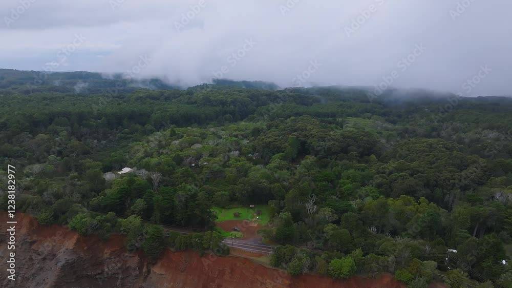 Aerial shot of a lush forest and red cliff on Kauai Island, Hawaii, with a road, vehicles, and a park. Slow sweeping motion under an overcast sky. Aerial shot of a lush forest and red cliff on Kauai Island, Hawaii, with a road, vehicles, and a park. Slow sweeping motion under an overcast sky.