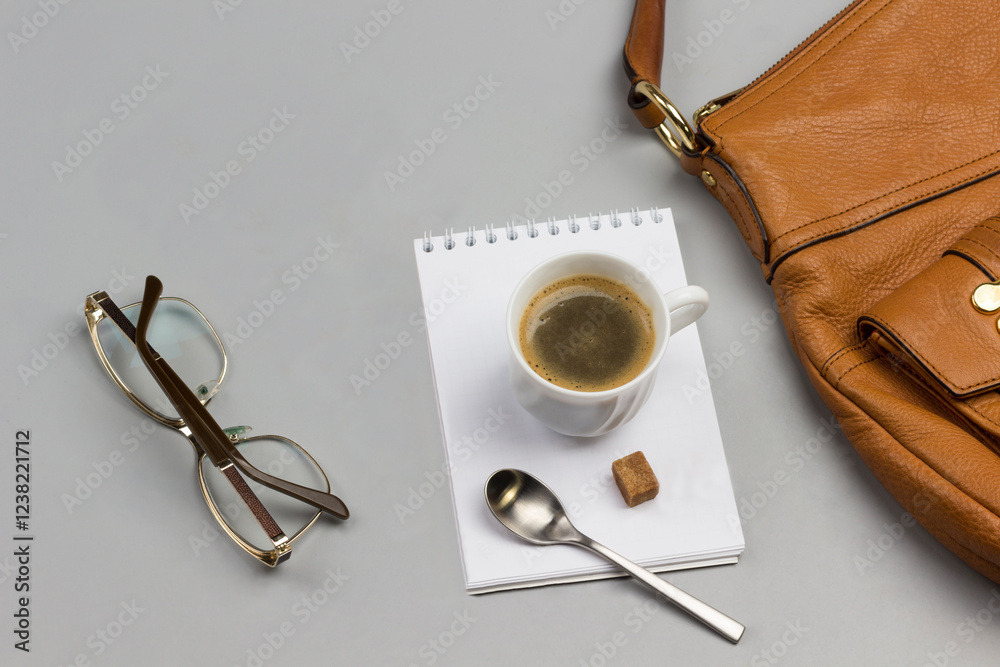 Cup of coffee, spoon and sugar cubes on notebook. Glasses, postcard and brown handbag on table