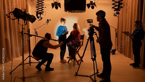 Studio for photo video production with lighting equipment and cyclorama, team members preparing lights and camera, stylist fixing model makeup before recording.