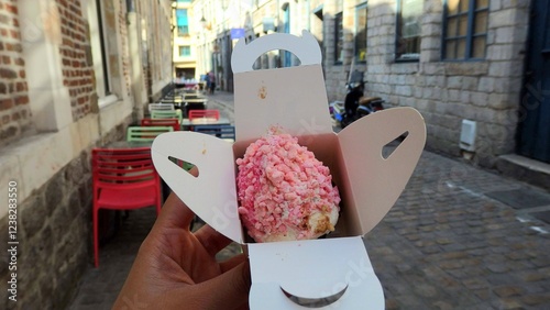 Hand holding Aux Merveilleux de Fred pink cake inside a paper packaging on the small alley outside the shop