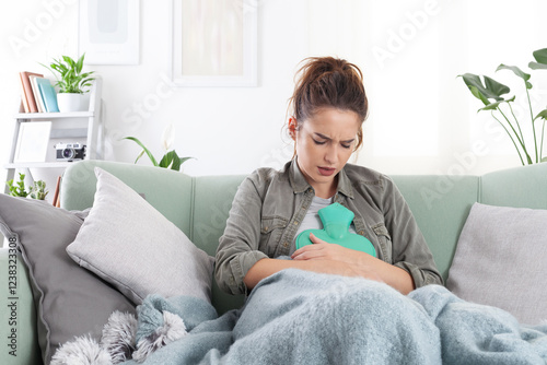 Young woman sitting on sofa at home suffering from period pain, cramps. Holding hot water bottle on abdomen, dealing with endometriosis, indigestion, and tummy pain