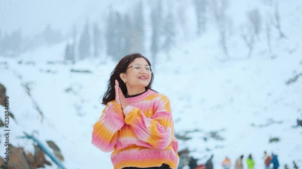 4K portrait of Indian teenager girl enjoying snowfall in mountains and rubbing hands due to cold. Tourist enjoying holidays at Pahalgam, Kashmir, India. Girl wearing multicolor warm sweater