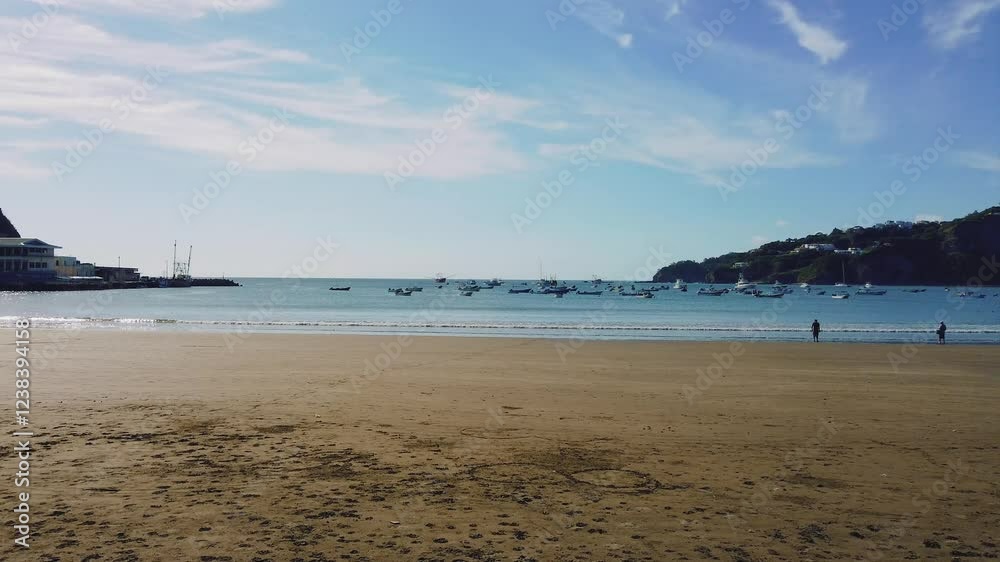 Sandy beach with calm ocean waves, fishing boats anchored in the water, and two people standing near the shore. Hills with houses in the background under a clear blue sky. Sandy beach with calm ocean waves, fishing boats anchored in the water, and two people standing near the shore. Hills with houses in the background under a clear blue sky.