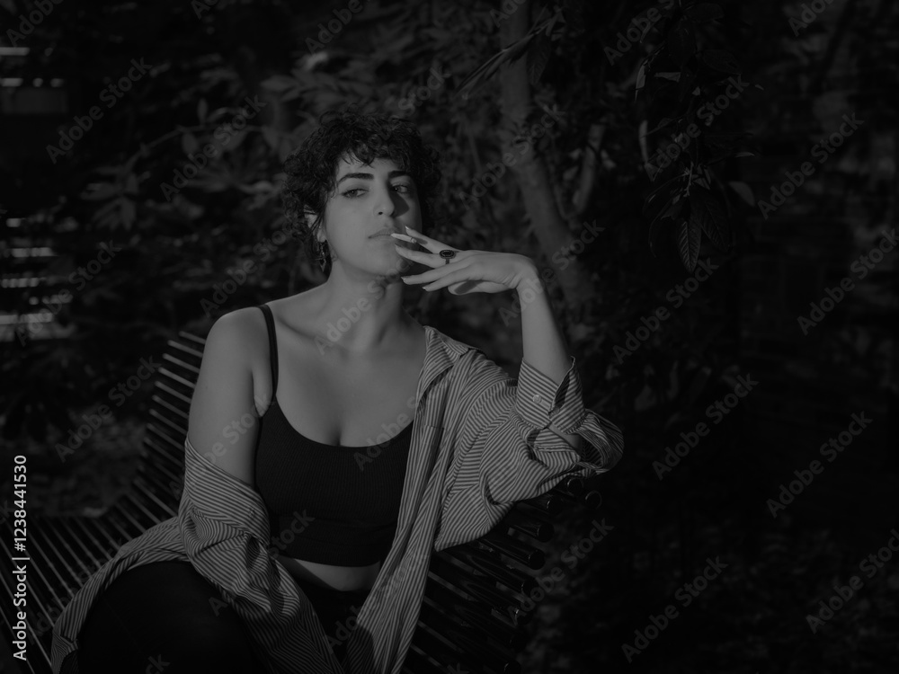 A young woman with curly hair sits on a bench at night, holding a cigarette with a pensive expression, captured in moody monochrome lighting A young woman with curly hair sits on a bench at night, holding a cigarette with a pensive expression, captured in moody monochrome lighting