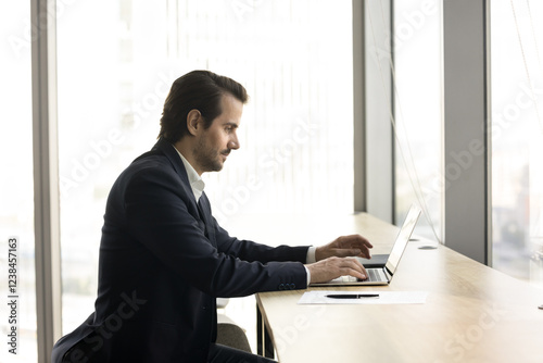 Young 35s businessman in suit typing on his laptop in workspace, looks focused, lead correspondence to client, send e-mail, solve business remotely, prepare report, work on presentation. Trading, tech photo