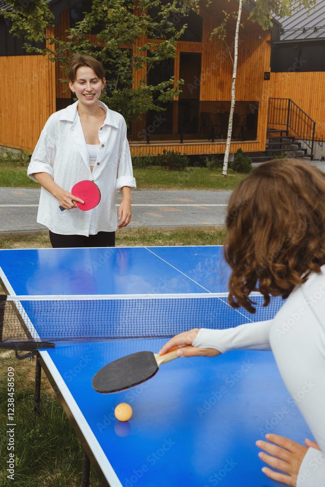 Obraz premium Two women engage in active game of ping pong at an outdoor table tennis setting. The area has green territory and wooden cabins, ideal summer outdoor leisure activities