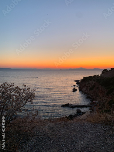 Coastal view at sunset with orange and blue gradient sky, calm sea, rugged cliffs, and sparse vegetation