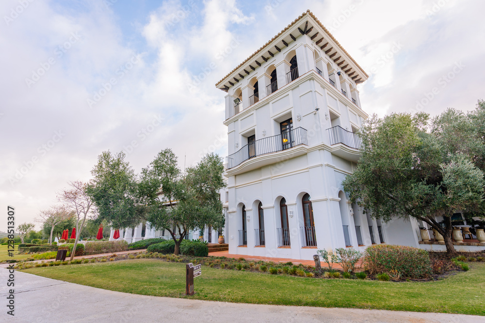 Obraz premium Elegant white Mediterranean-style building with arched windows, balconies, lush greenery, and a manicured lawn under a partly cloudy sky..