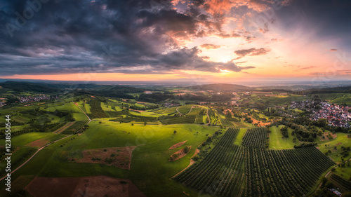 A magnificent aerial view of rolling vineyards and lush fields under a glowing sunset sky. The landscape features a small village, adding to the charm of this serene countryside scene