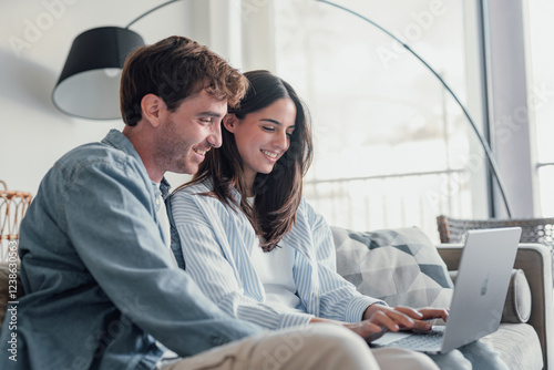 Happy millennial couple sit relax on couch in living room watching video on laptop together, smiling young husband and wife rest on sofa at home browsing Internet using modern computer device.