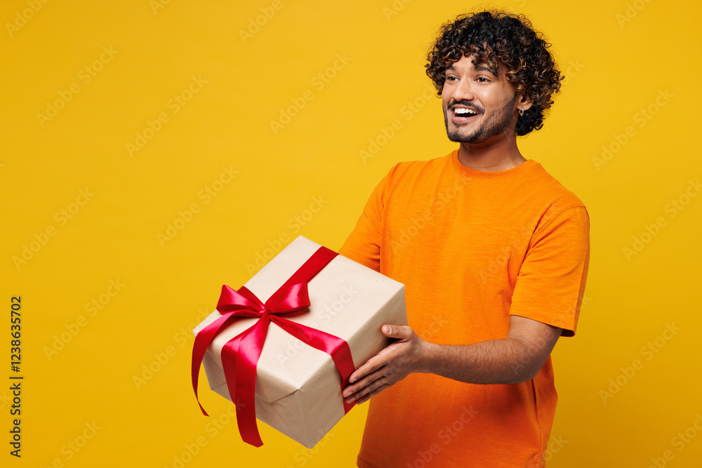 Side view young smiling fun happy Indian man he wear orange t-shirt casual clothes hold in hand present box with gift ribbon bow isolated on plain yellow background studio portrait. Lifestyle concept. Side view young smiling fun happy Indian man he wear orange t-shirt casual clothes hold in hand present box with gift ribbon bow isolated on plain yellow background studio portrait. Lifestyle concept.