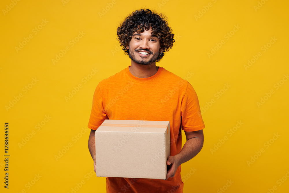 Fototapeta premium Young smiling cheerful fun cool happy Indian man he wear orange t-shirt casual clothes hold blank cardboard box looking camera isolated on plain yellow background studio portrait. Lifestyle concept.