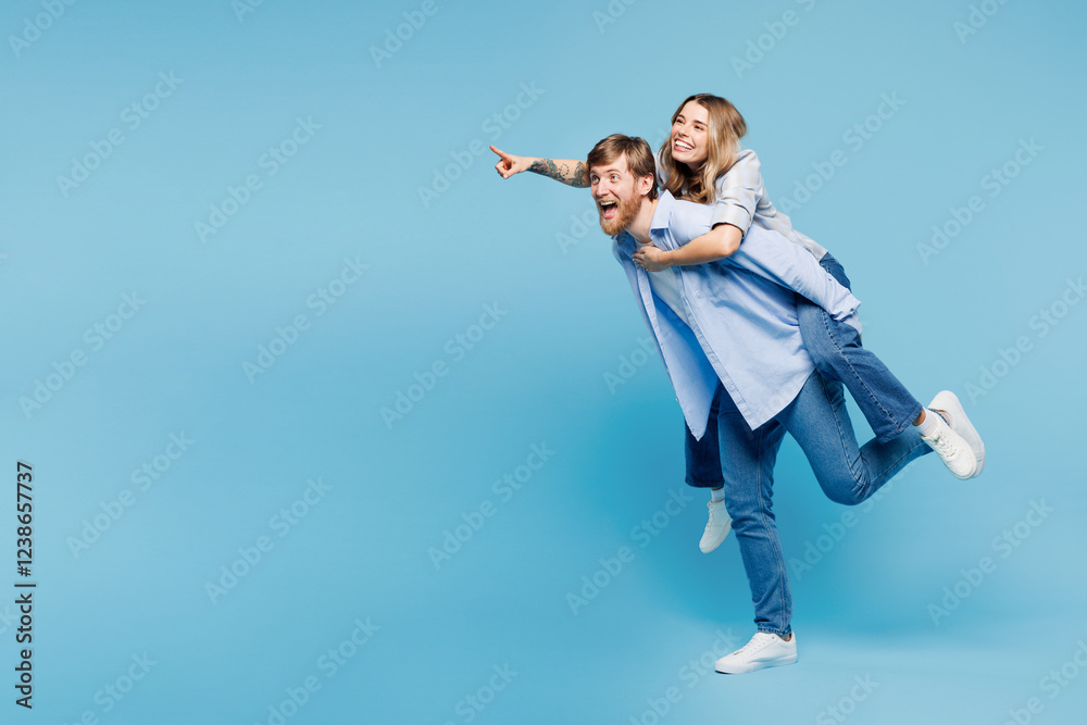Full body young couple two friends family man woman wearing shirt casual clothes together giving piggyback ride to joyful, sit on back point finger aside run isolated on pastel plain blue background. Full body young couple two friends family man woman wearing shirt casual clothes together giving piggyback ride to joyful, sit on back point finger aside run isolated on pastel plain blue background.