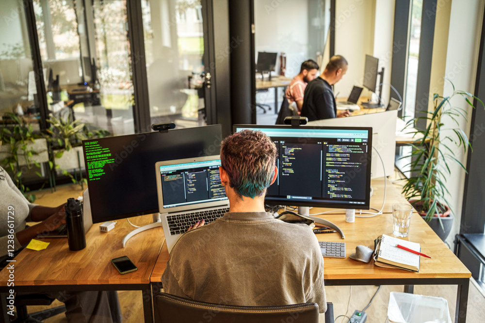 High angle rear view of non-binary coding professional working on desktop PC and laptop in office High angle rear view of non-binary coding professional working on desktop PC and laptop in office