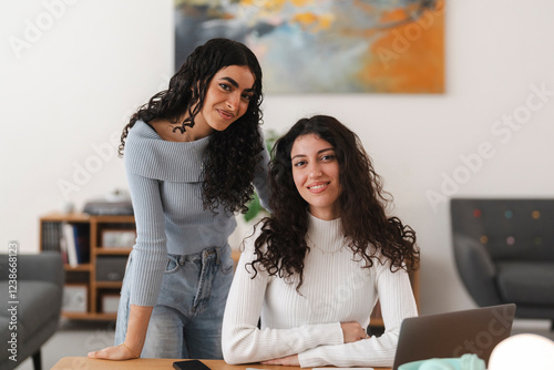 Two young women with curly hair are posing together at a wooden desk in a bright and modern workspace, smiling with laptops, notebooks, and office supplies around them. photo