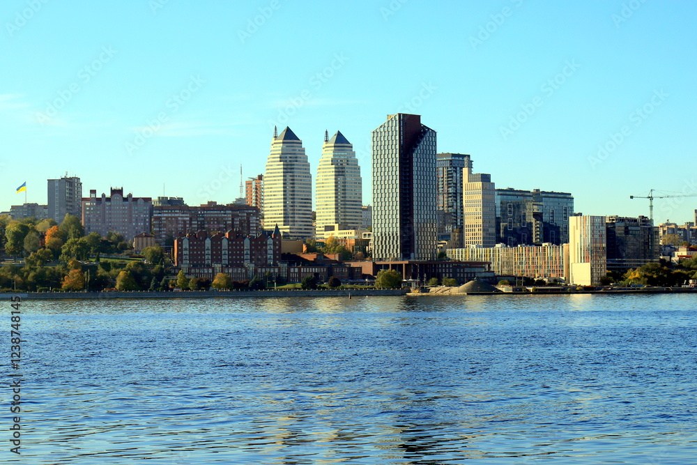 Obraz premium Big city on banks wide river. Ukrainian city Dnipro, Ukraine. Beautiful modern towers, buildings, skyscrapers are reflected in water morning, river flows, Dnepr