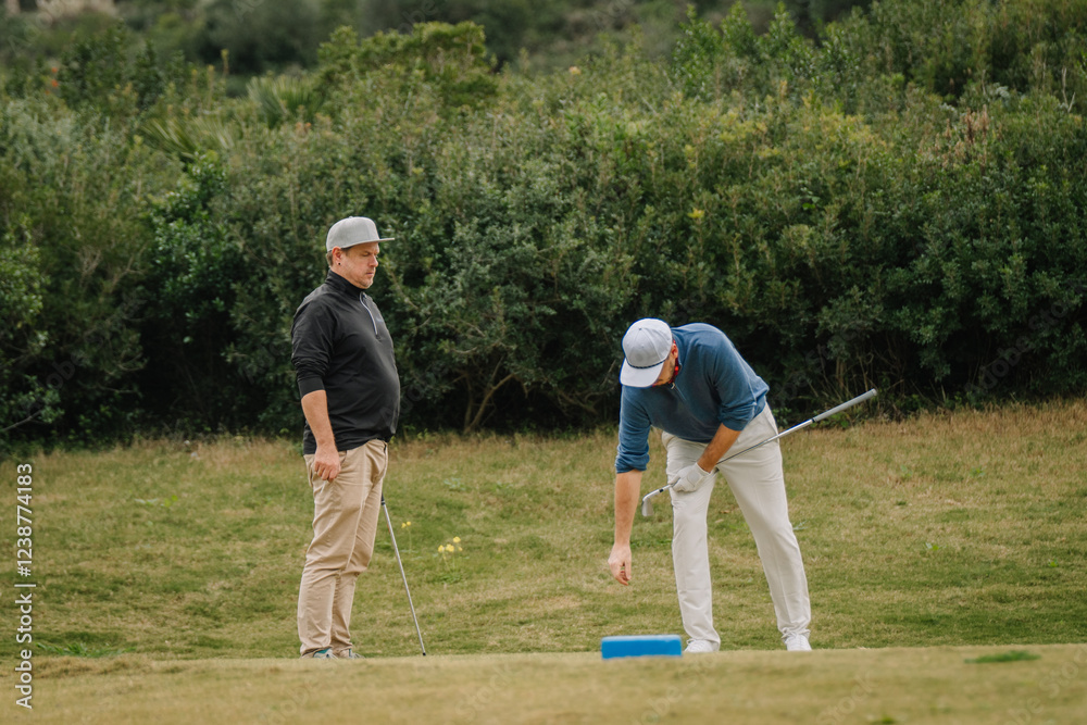 Two golfers on a grassy course, one placing the ball while the other observes, both holding golf clubs, surrounded by greenery. Two golfers on a grassy course, one placing the ball while the other observes, both holding golf clubs, surrounded by greenery.