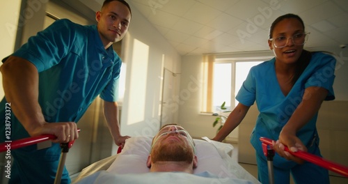 A man with an oxygen tube in his nose is lying on a medical gurney. The orderlies talk and smile as they take the patient through the hospital corridor