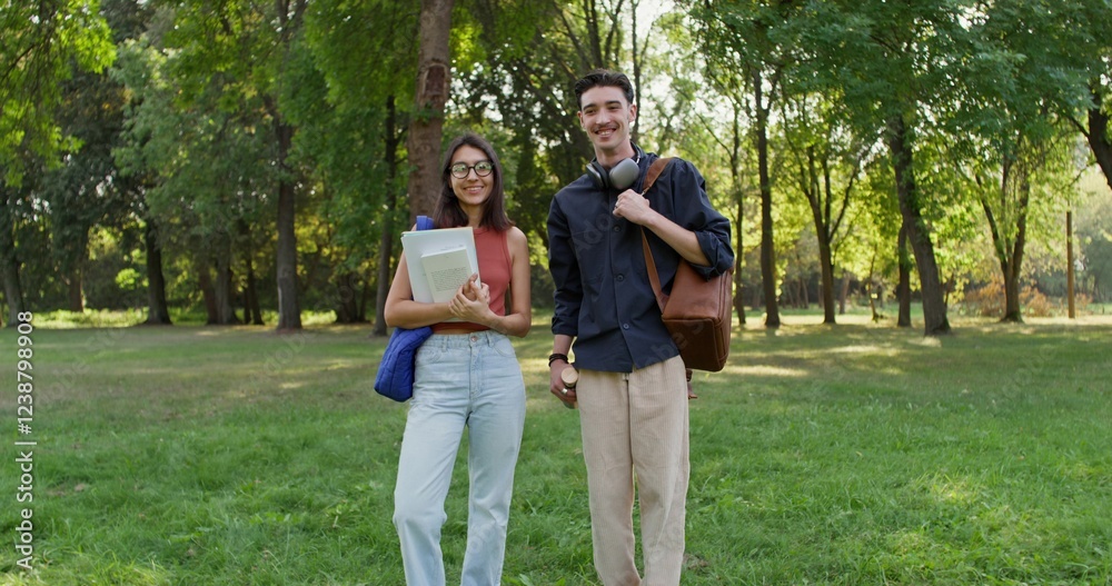 Fototapeta premium A young man and a woman with backpacks and notebooks in their hands are smiling at the camera. They are standing in the city park on a summer day