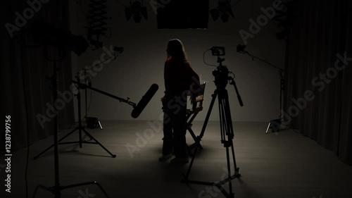 Studio for photo video production with lighting equipment and cyclorama, model actress sits on director chair in the middle under spotlight in front of camera.