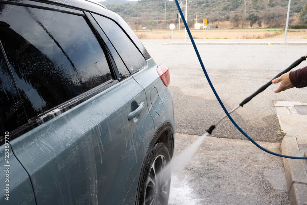 man cleaning car at pressure washing site with hose, light blue car. Detail of the water pressure and the back of the car.