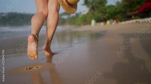 Barefoot woman walks on sandy shoreline holding straw hat, feet sinking into wet sand by sea. Leisure stroll along tropical beach, feeling texture of waves on skin, person enjoying vacation freedom.