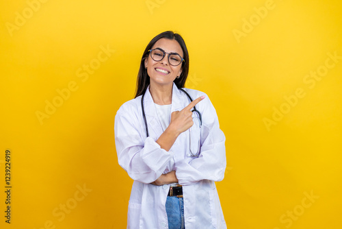 Young brunette doctor woman wearing stethoscope standing over isolated yellow background smiling and pointing with hand and finger to the side