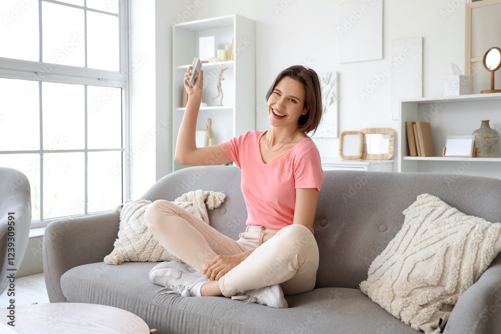 Winking young woman with air conditioner remote control sitting on sofa in living room Winking young woman with air conditioner remote control sitting on sofa in living room