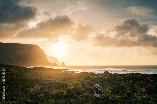 Sun ascends behind Motu Marotiri Stake and Cape Roggeveen, located on the southern side of the Poike Peninsula in Rapa Nui (Easter Island), Chile. A beach filled with black rocks in the foreground. photo